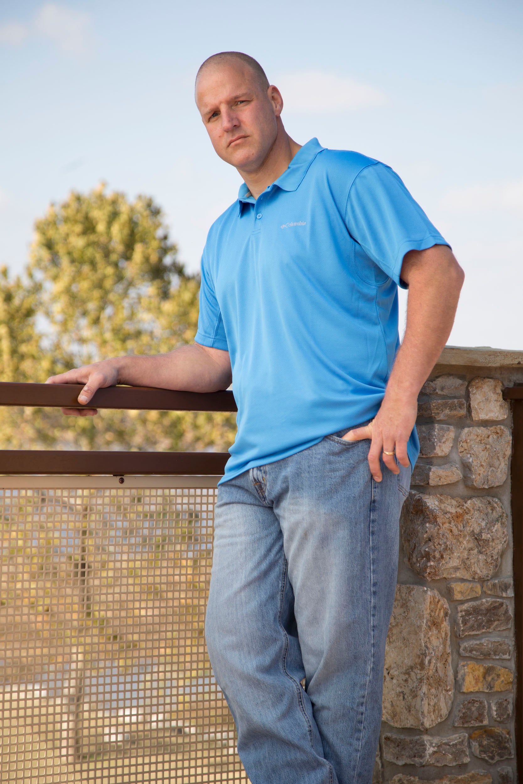 Man wearing a blue polo shirt and jeans with a concealed holster around his waist standing on a balcony with a stone wall and trees in the background.