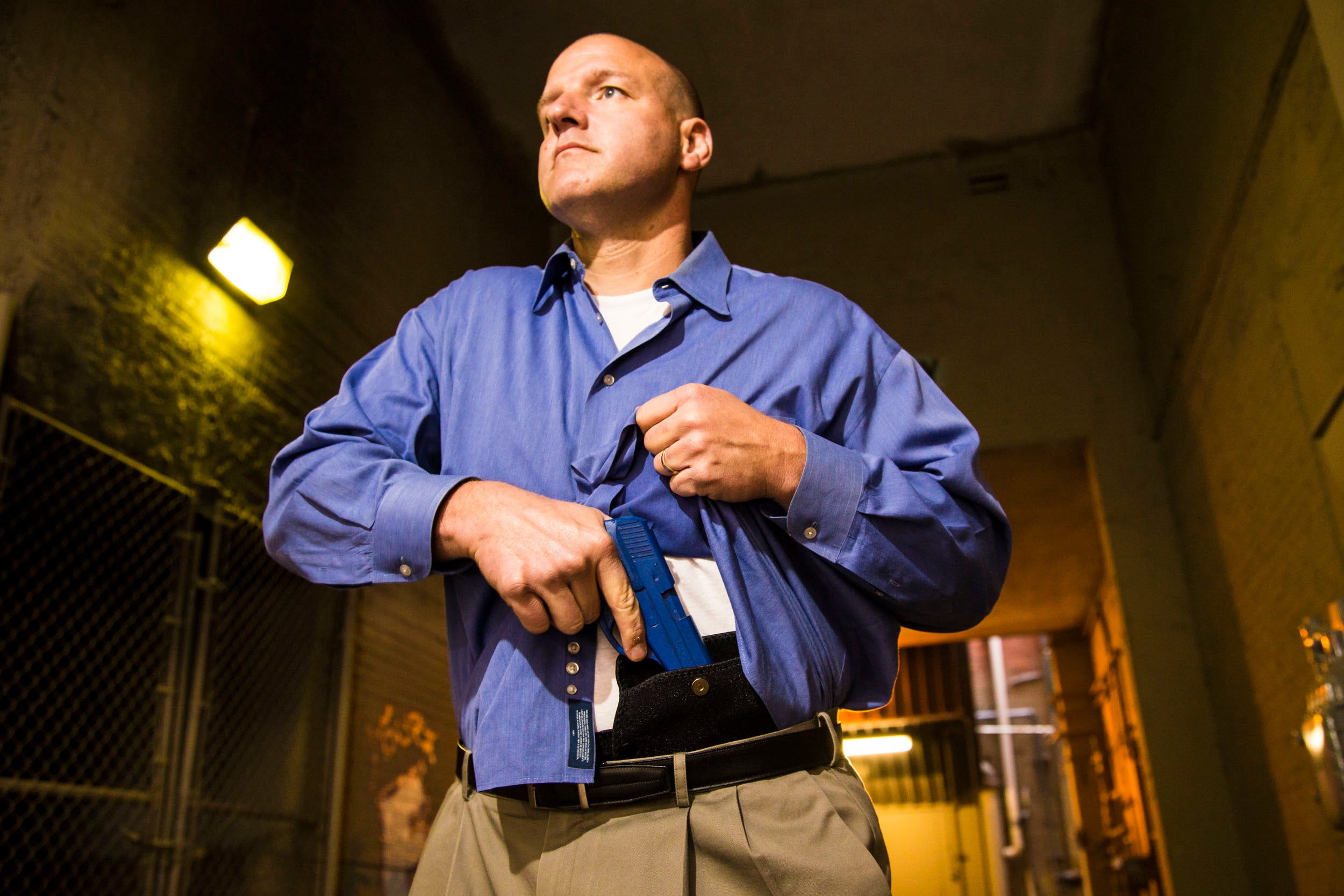 Man in a blue shirt and khaki pants standing in an indoor setting wearing a holster