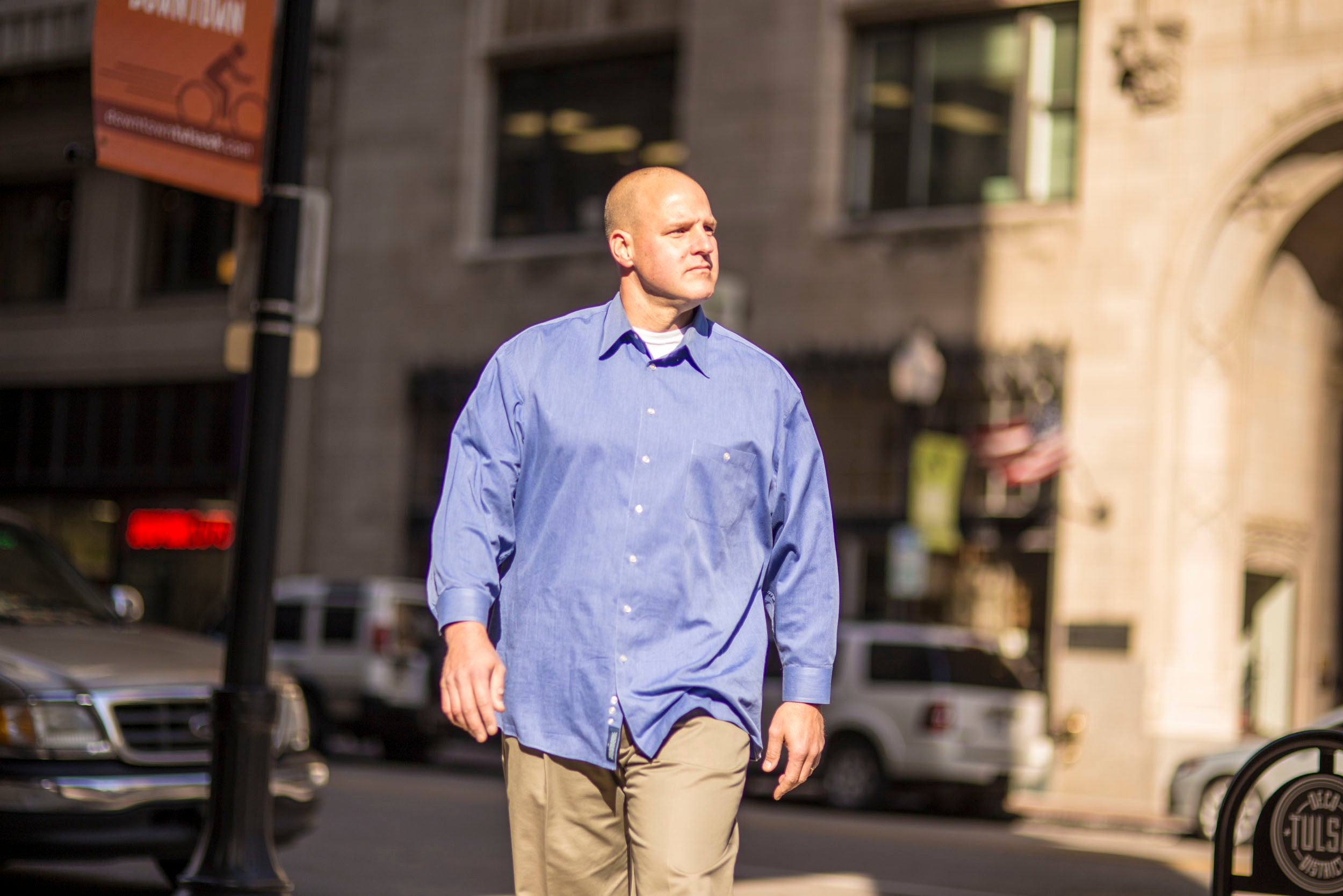Man in a blue shirt walking on a city street with buildings and vehicles in the background