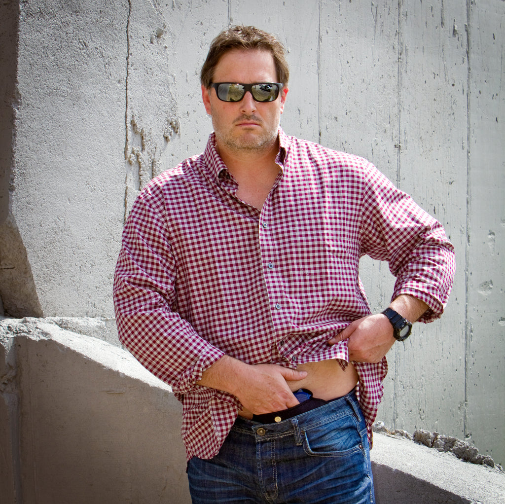 Man wearing a red and white checkered shirt and sunglasses, leaning against a concrete wall wearing concealed holster