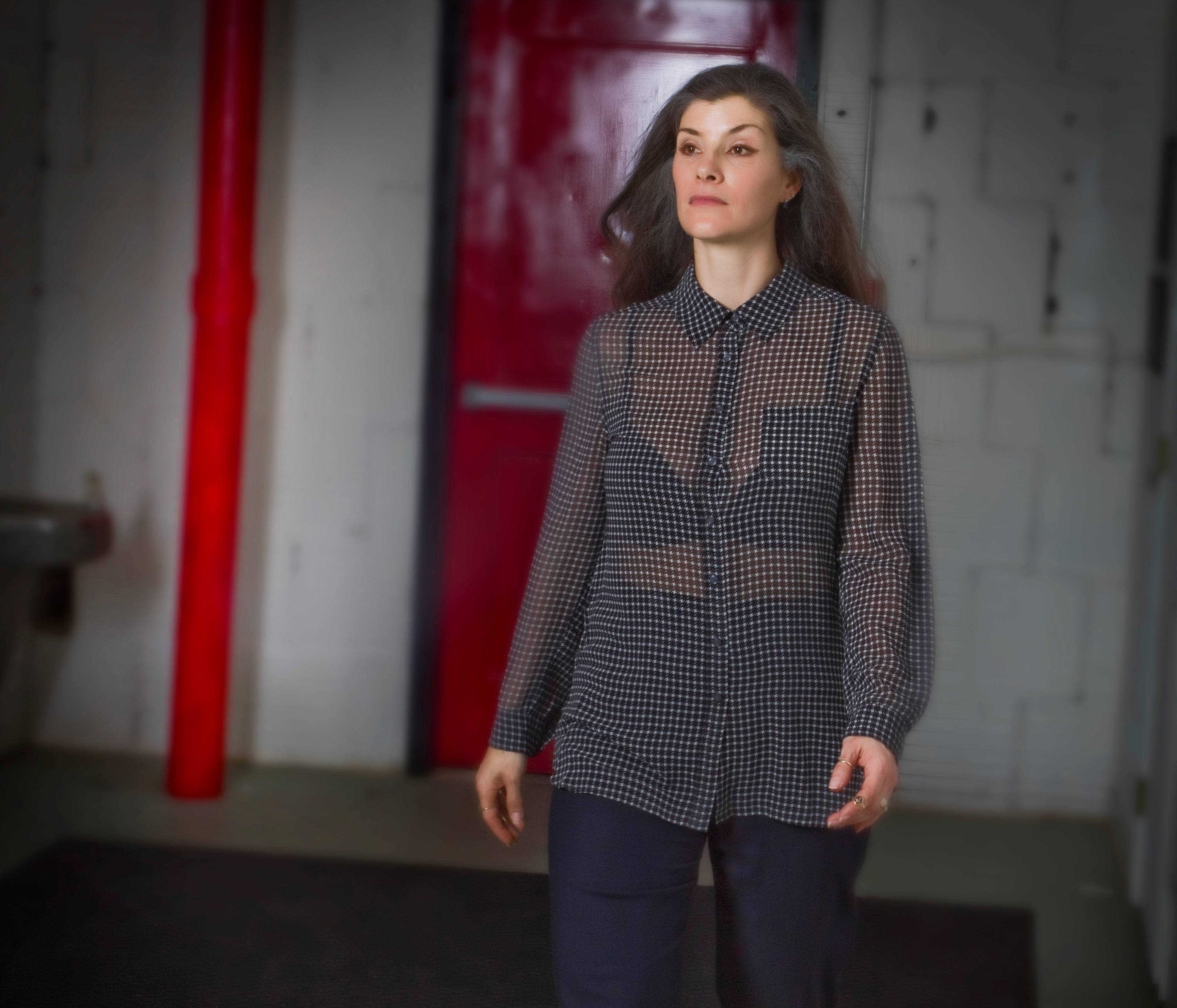Woman wearing a checkered shirt in an industrial setting with red pipes and white walls carrying concealed firearm